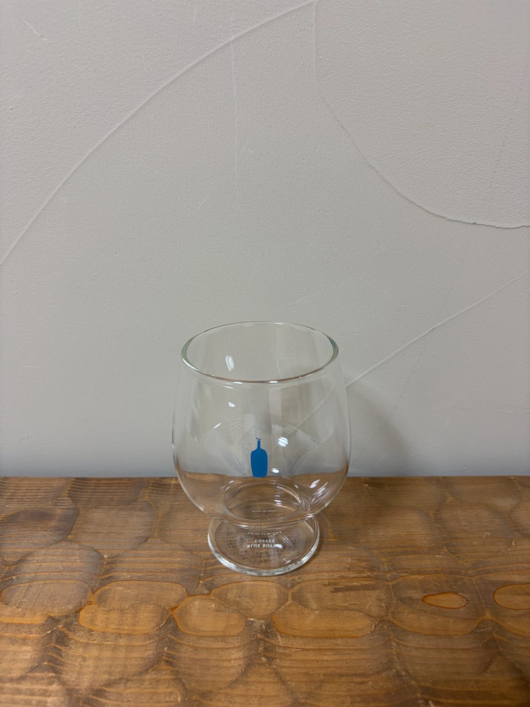 木の台に置いた空のゴブレットグラス。胴に青いボトルのロゴ、脚にBLUE BOTTLE COFFEEの文字が入っている